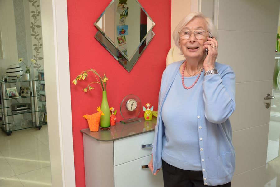 Eine Seniorin steht im Flur und hat ein Telefon in der Hand. 