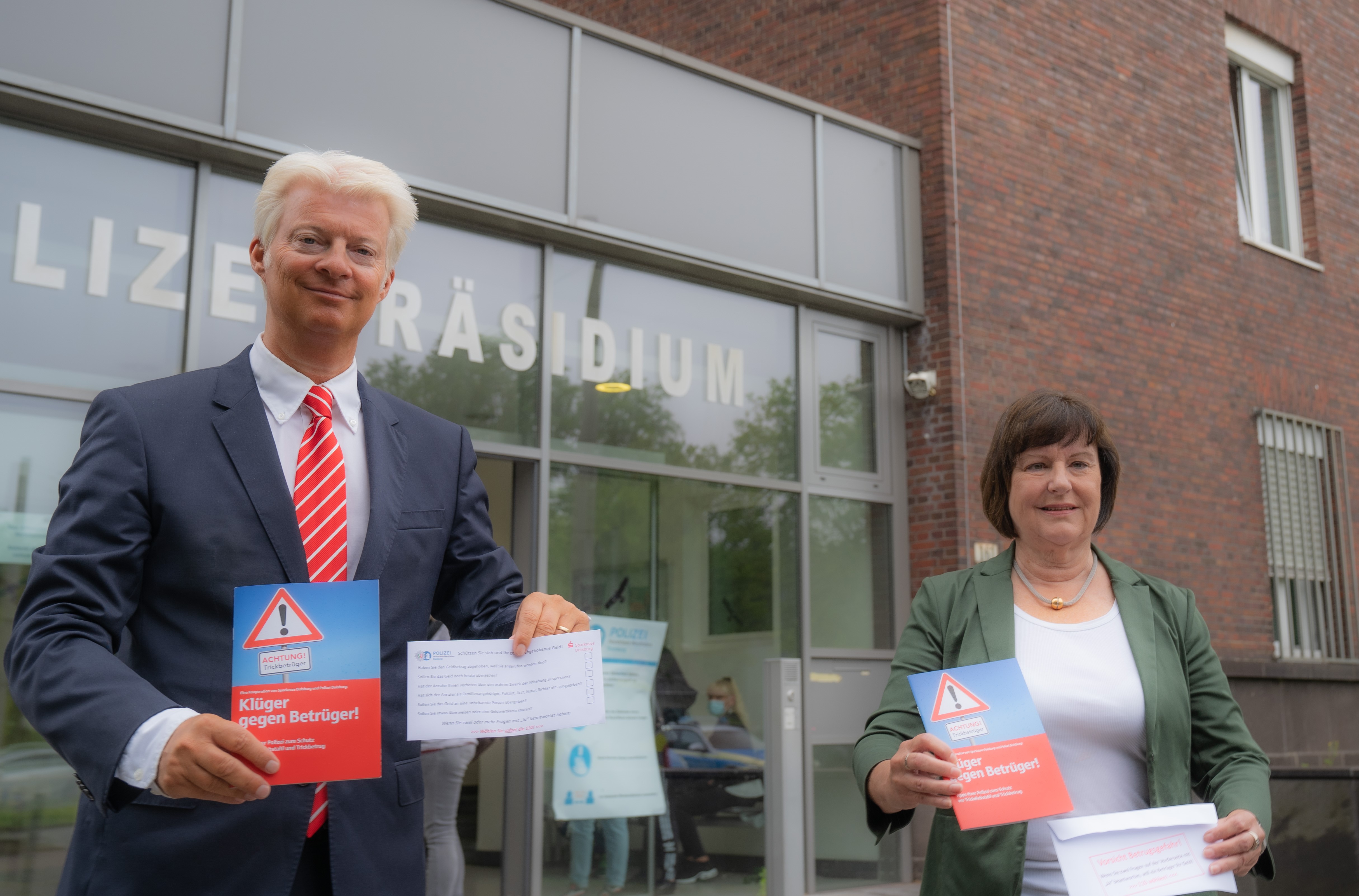  Dr. Joachim Bonn und Dr. Elke Bartels mit der Broschüre "Klüger gegen Betrüger" vor dem Polizeipräsidium Duisburg